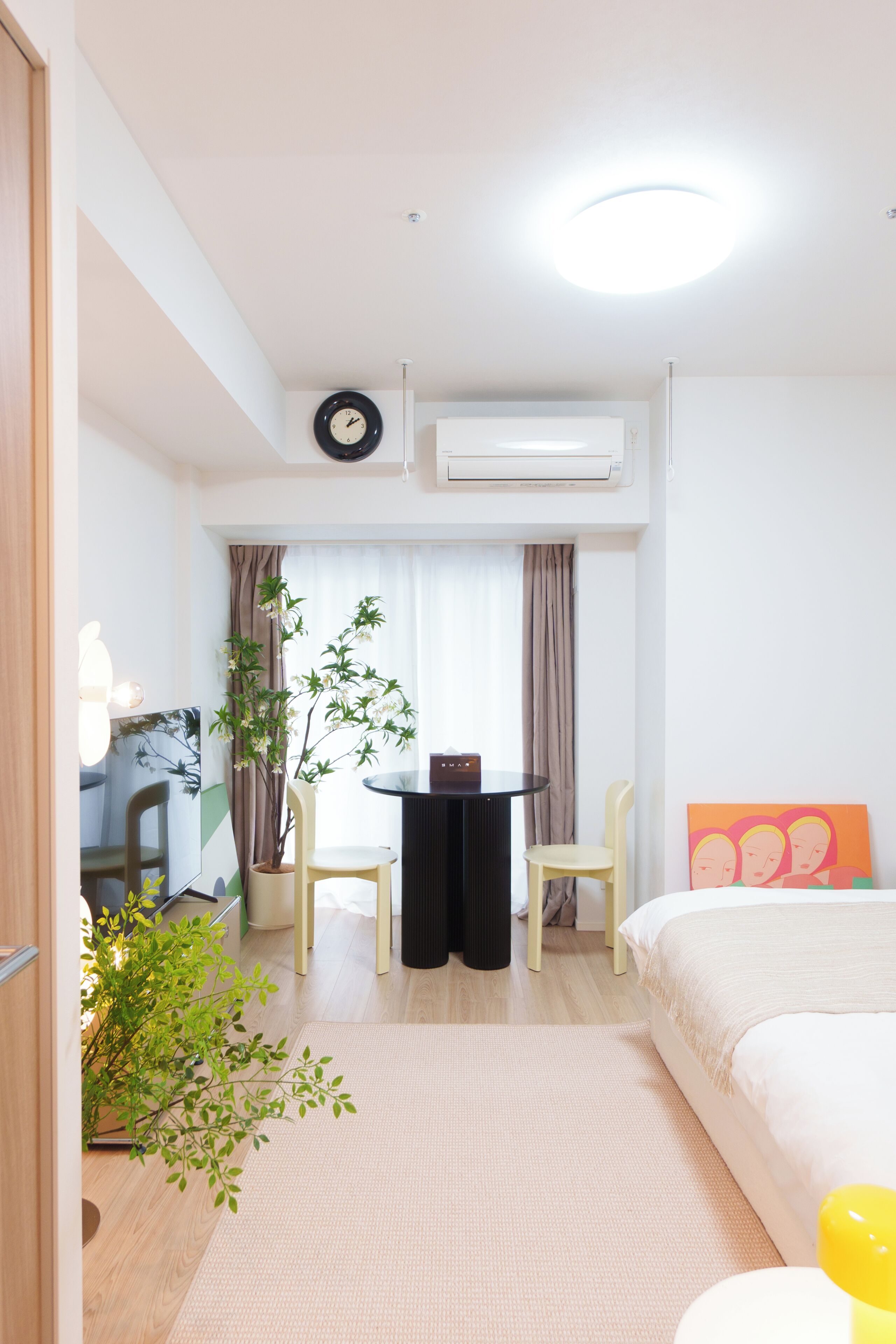 A hotel room with a bed, a desk, a chair, a potted plant, and a window with curtains.