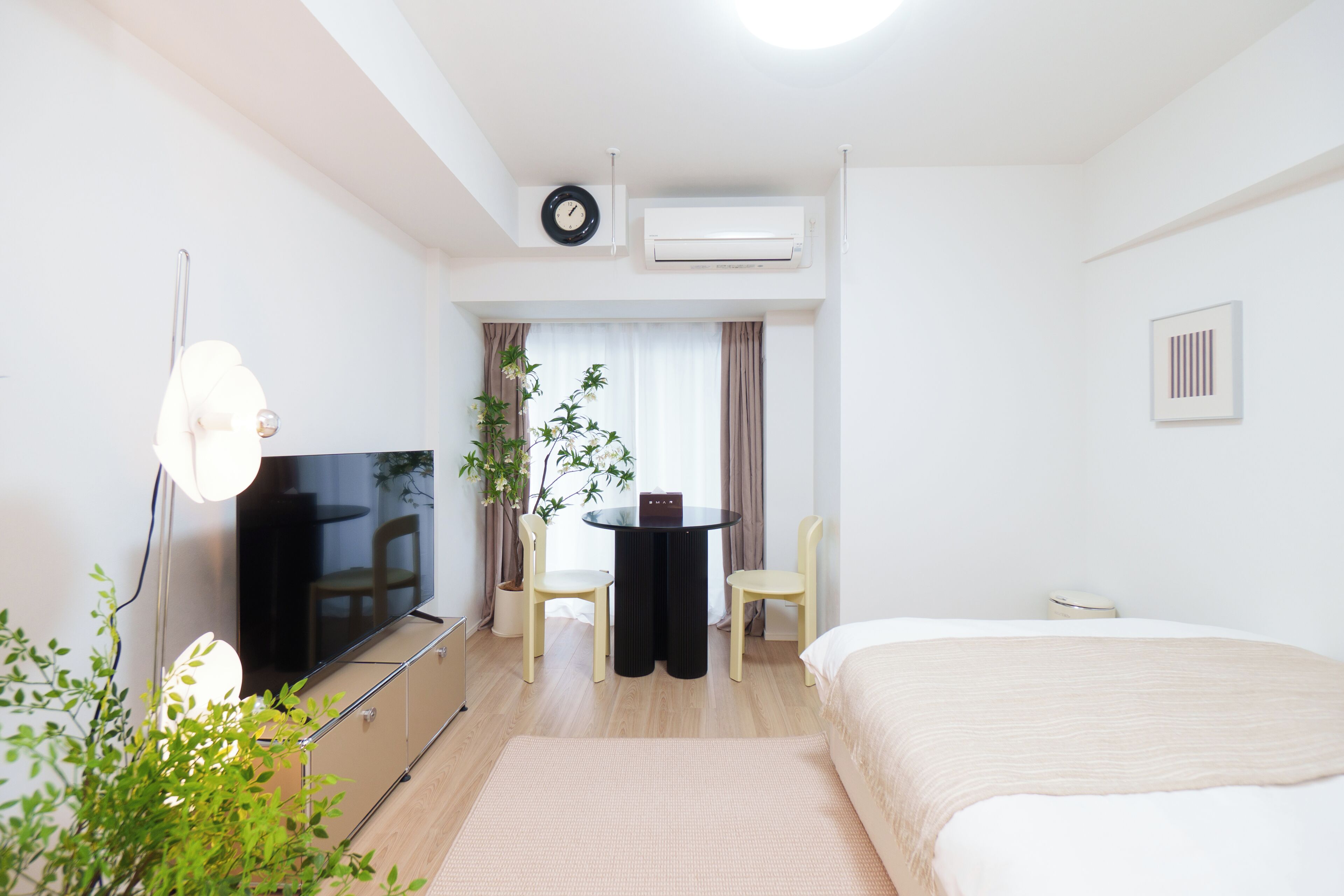 A hotel room with a bed, a TV, a desk with two chairs, a potted plant, and a window with curtains.