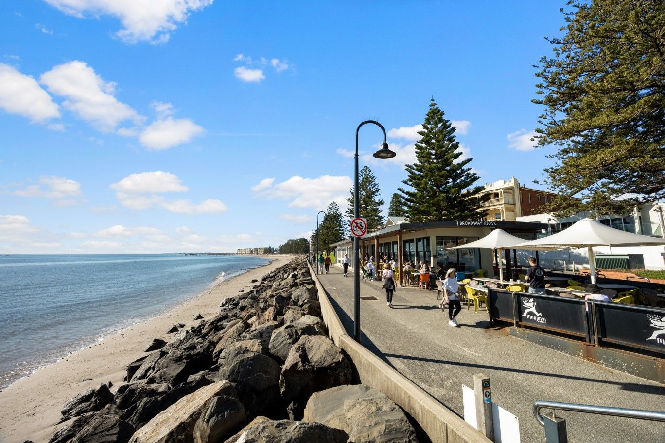 A coastal promenade with a rocky shoreline, a pedestrian path, and outdoor seating areas.
