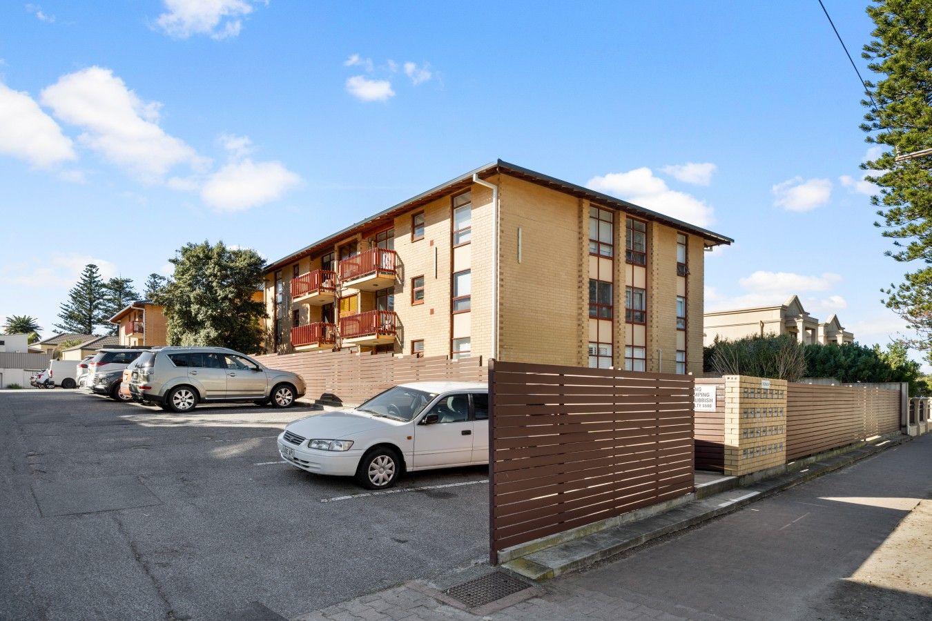 Un edifici residencial de diverses plantes amb una entrada tancada per reixa, cotxes aparcats i un cel serè.