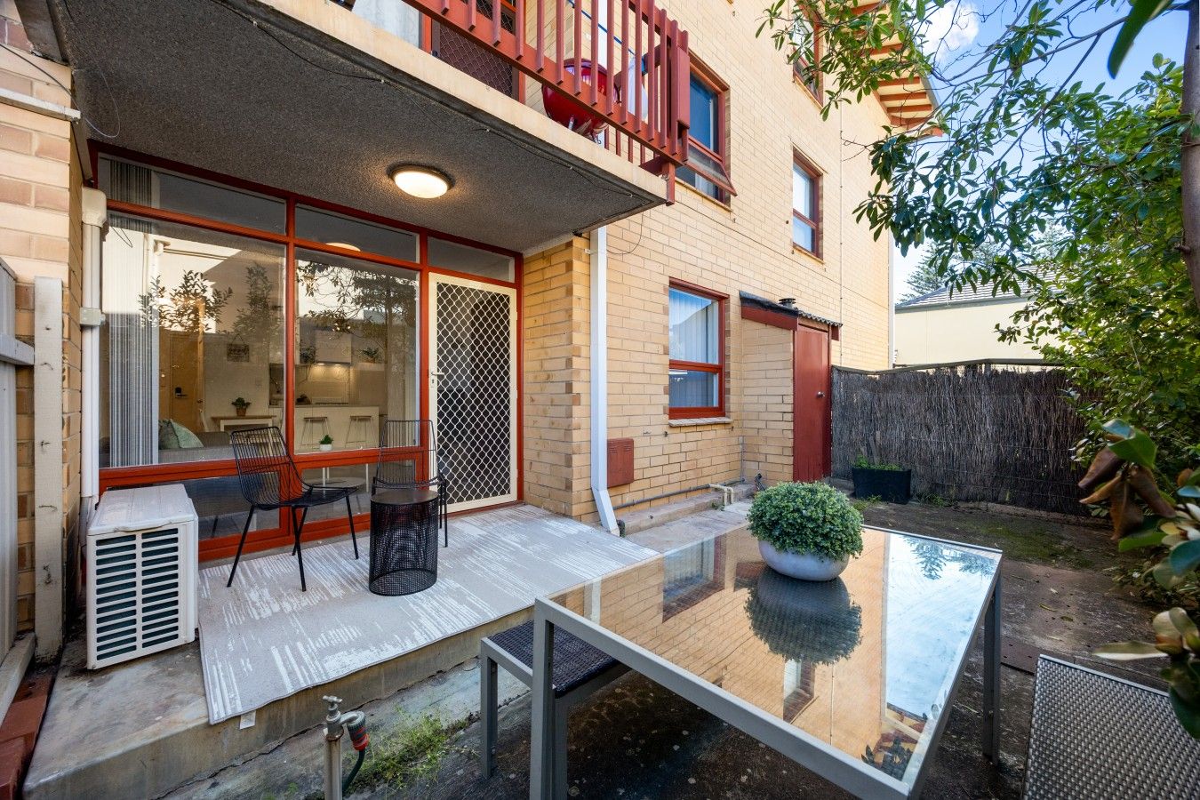 A modern patio with a glass table and chairs, a potted plant, and a red balcony railing.