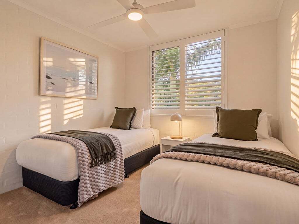 Ein Schlafzimmer mit zwei Betten, einem Deckenventilator, einem Fenster mit Jalousien und einer Nachttischlampe.