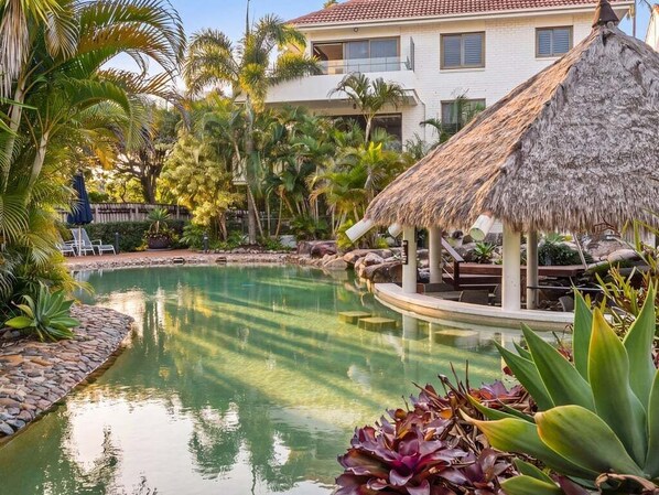 A pool area with a thatched structure, surrounded by tropical plants and a white building in the background.