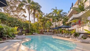 A swimming pool with lounge chairs and umbrellas, surrounded by palm trees and a building with balconies.