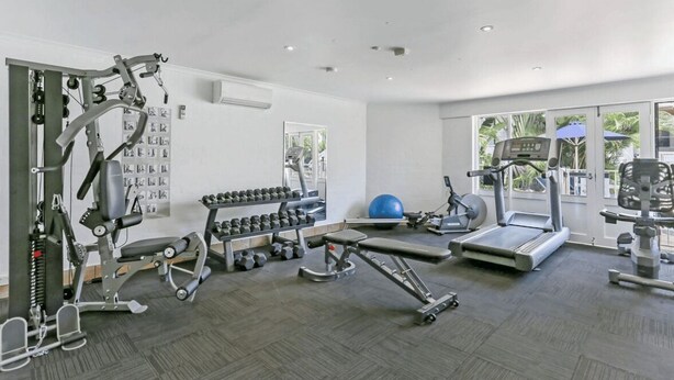 A gym with various exercise equipment, a wall-mounted rack of dumbbells, and a large window providing natural light.