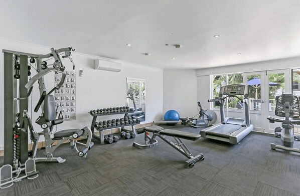A gym with various exercise equipment, a wall-mounted rack of dumbbells, and a large window providing natural light.