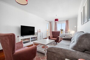 Ein modernes Wohnzimmer mit Flachbildfernseher, einer Couch, zwei Sesseln, einem Couchtisch und einem Essbereich mit Tisch und Stühlen.