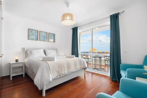 Een slaapkamer met een bed, nachtkastje, lamp en uitzicht op de zee door een groot raam.