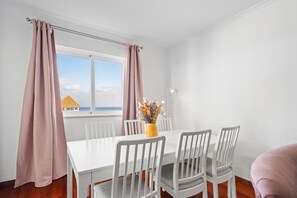 Ein Essbereich mit einem weißen Tisch und Stühlen, einer Vase mit Blumen und einem Fenster mit Vorhängen.