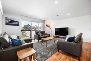 Ein modernes Wohnzimmer mit einer Couch, einem Sessel und einem Couchtisch, einem Flachbildfernseher und Blick ins Freie.