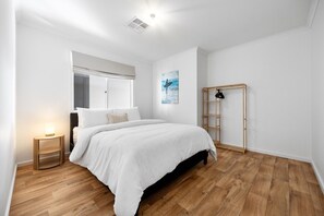 Et soveværelse med en seng, et sengebord med lampe, et vindue med persienner, et billede på væggen og en træstige lænet mod væggen.