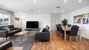 A modern living room with a flat-screen TV, a sofa, a dining table with chairs, and a large window. - A Swell Time—Spacious Garden Haven by the Coast (Goolwa Beach)
