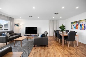 A modern living room with a flat-screen TV, a sofa, a dining table with chairs, and a large window. - A Swell Time—Spacious Garden Haven by the Coast (Goolwa Beach)