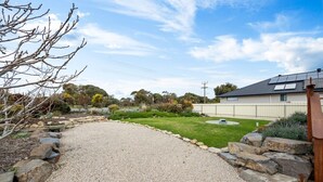 A gravel driveway leading to a house with a solar panel roof, surrounded by a landscaped garden with a lawn and various shrubs. - A Swell Time—Spacious Garden Haven by the Coast (Goolwa Beach)