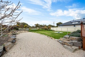 A gravel driveway leading to a house with a solar panel roof, surrounded by a landscaped garden with a lawn and various shrubs. - A Swell Time—Spacious Garden Haven by the Coast (Goolwa Beach)