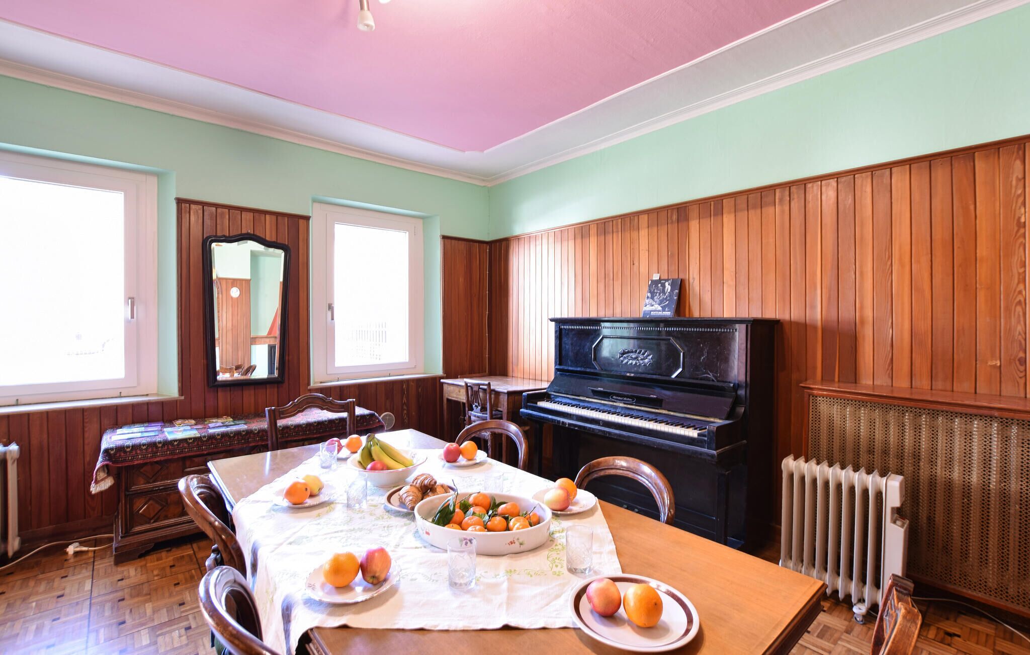 Ruokailuhuone, jossa on piano, puupanelointi, hedelmillä katettu pöytä ja suuri peili.