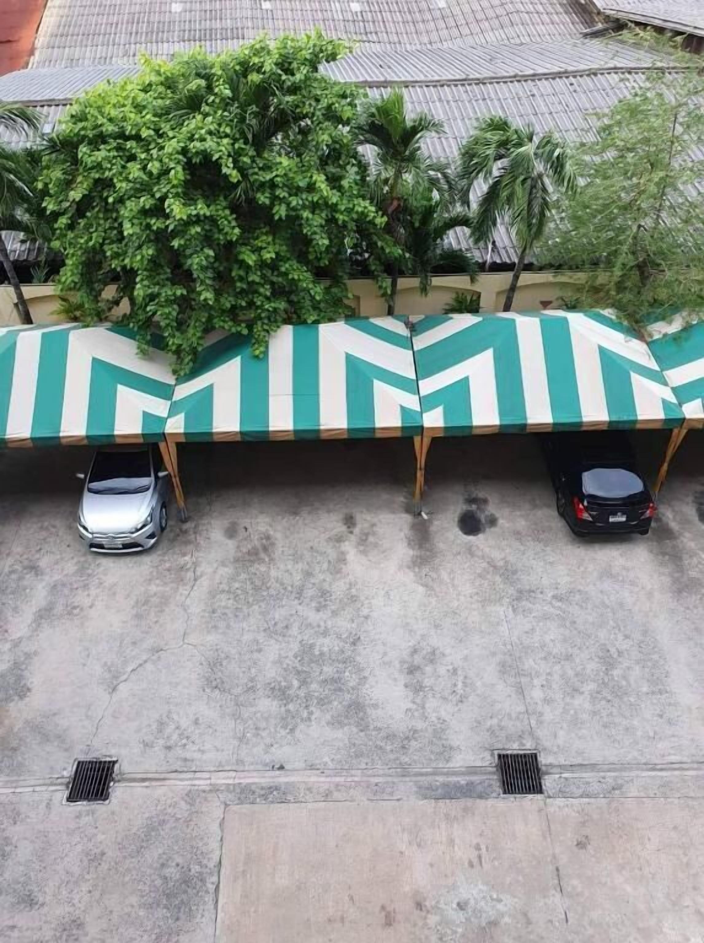 A parking area with two cars under striped awnings, a tree, and a building with a tiled roof.