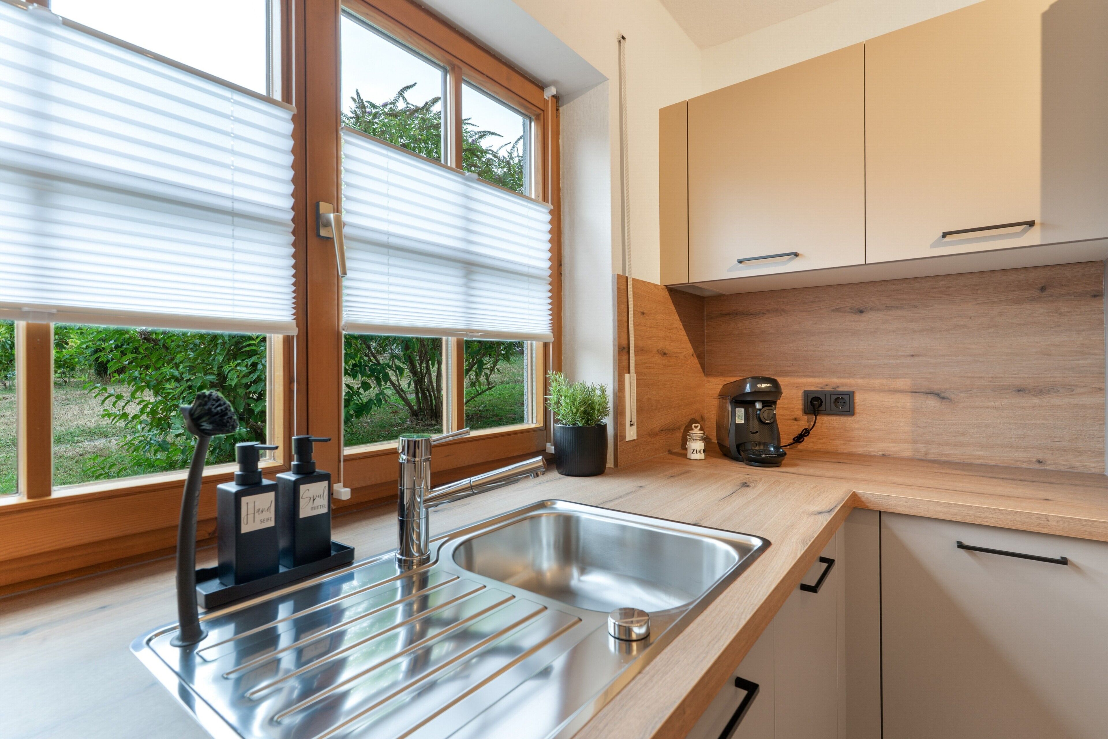 Une cuisine moderne dotée d’un évier en acier inoxydable, de meubles en bois et d’une fenêtre avec des stores.