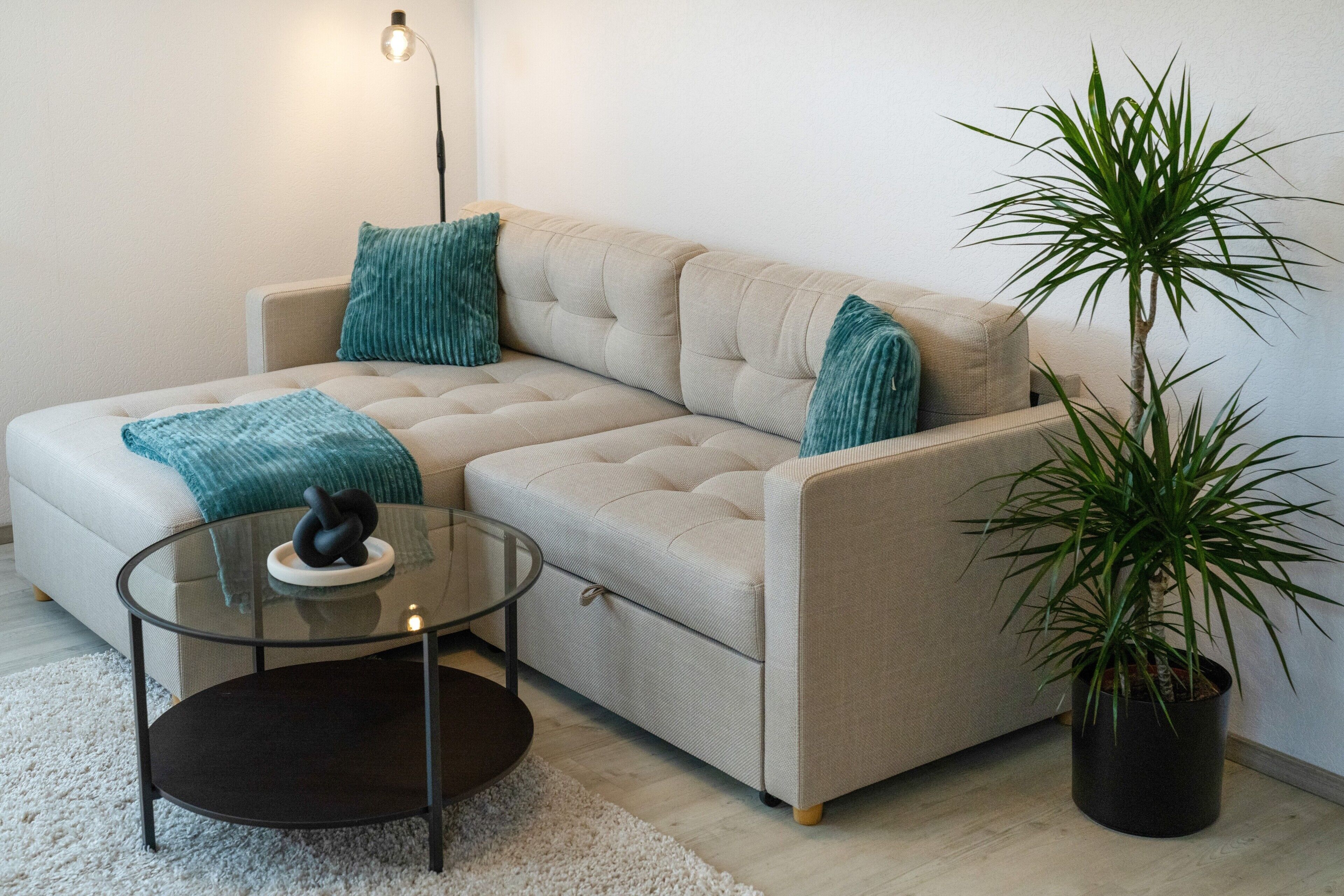 Ein beiges Ecksofa mit zwei türkisfarbenen Zierkissen, einem Glastisch mit einer schwarzen Skulptur und einer Topfpflanze.