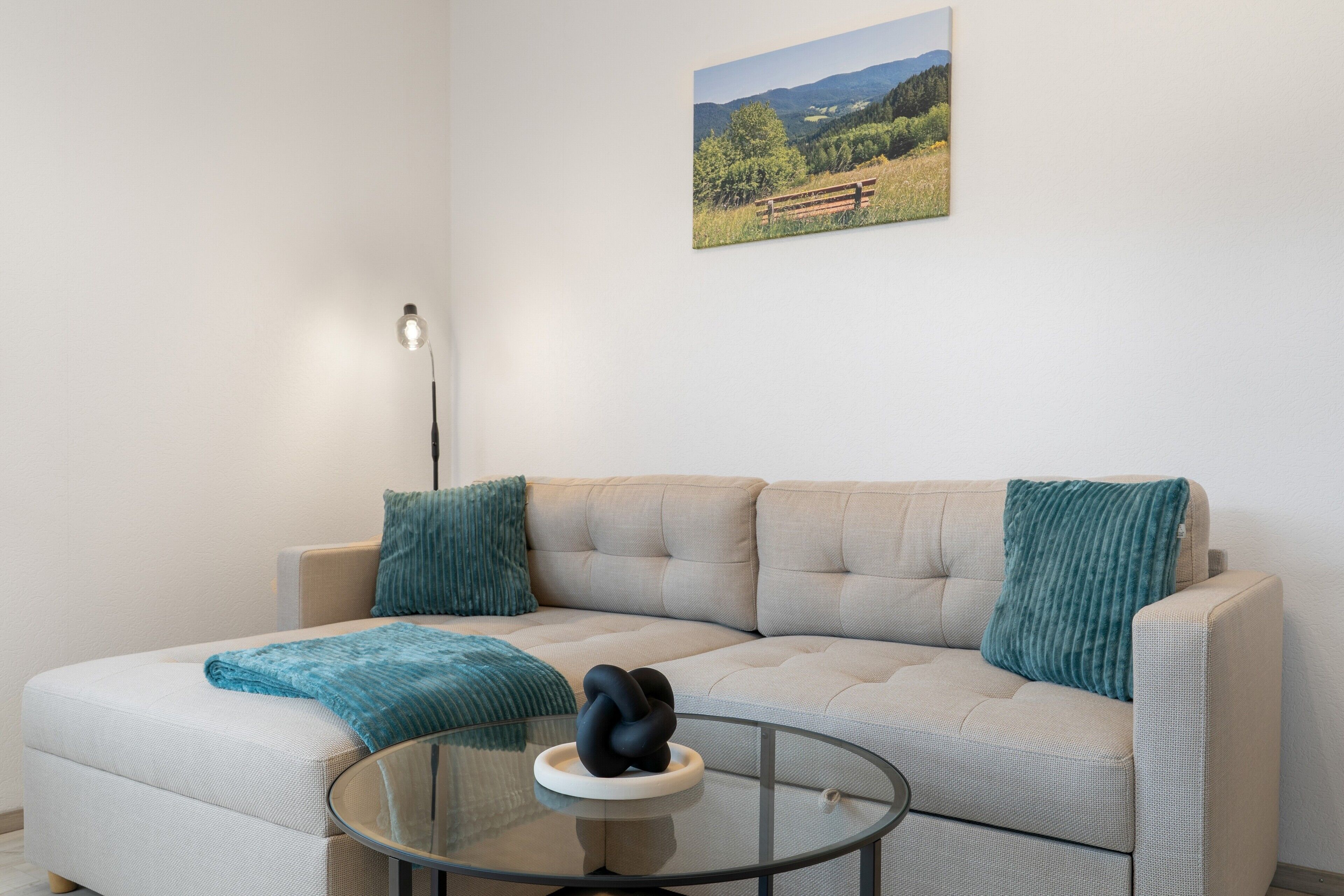 Ein Wohnzimmer mit einem beigefarbenen Sofa, zwei türkisfarbenen Kissen, einem Glastisch mit einer schwarzen Skulptur, einer Stehlampe und einem Landschaftsbild an der Wand.