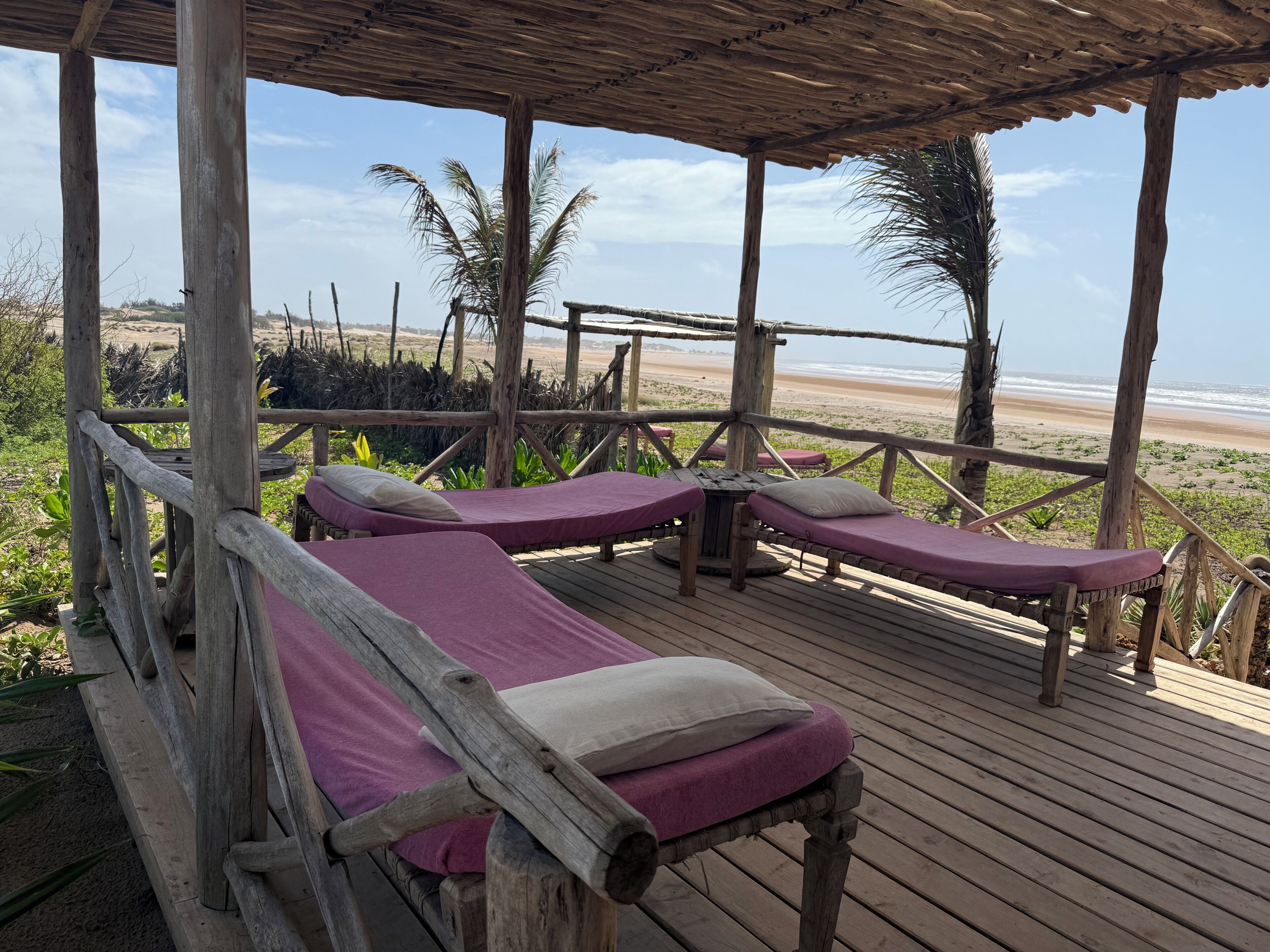 Eine Holzterrasse mit zwei Liegen, jeweils mit Kopfkissen und Decke, mit Blick auf einen Strand.