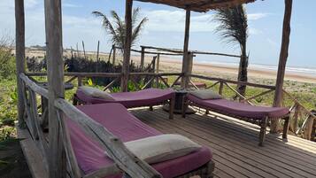 Eine Holzterrasse mit zwei Liegen, jeweils mit Kopfkissen und Decke, mit Blick auf einen Strand.