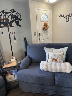 A living room with a gray sofa, a decorative wall piece, a door with a wreath, and a small table with a sign. - Beautiful, cozy 3 bedroom, two bath, private home on acreage in the mountains (Lava Hot Springs)