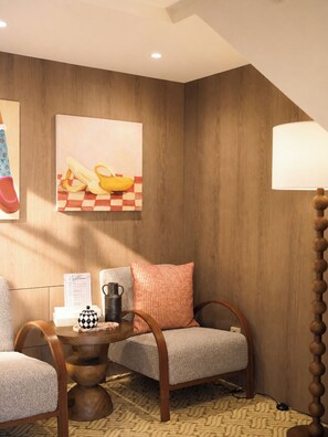 Una habitación acogedora con suelo a cuadros, pared de madera, un cuadro de plátanos, una lámpara, una mesa con una jarra y una silla.
