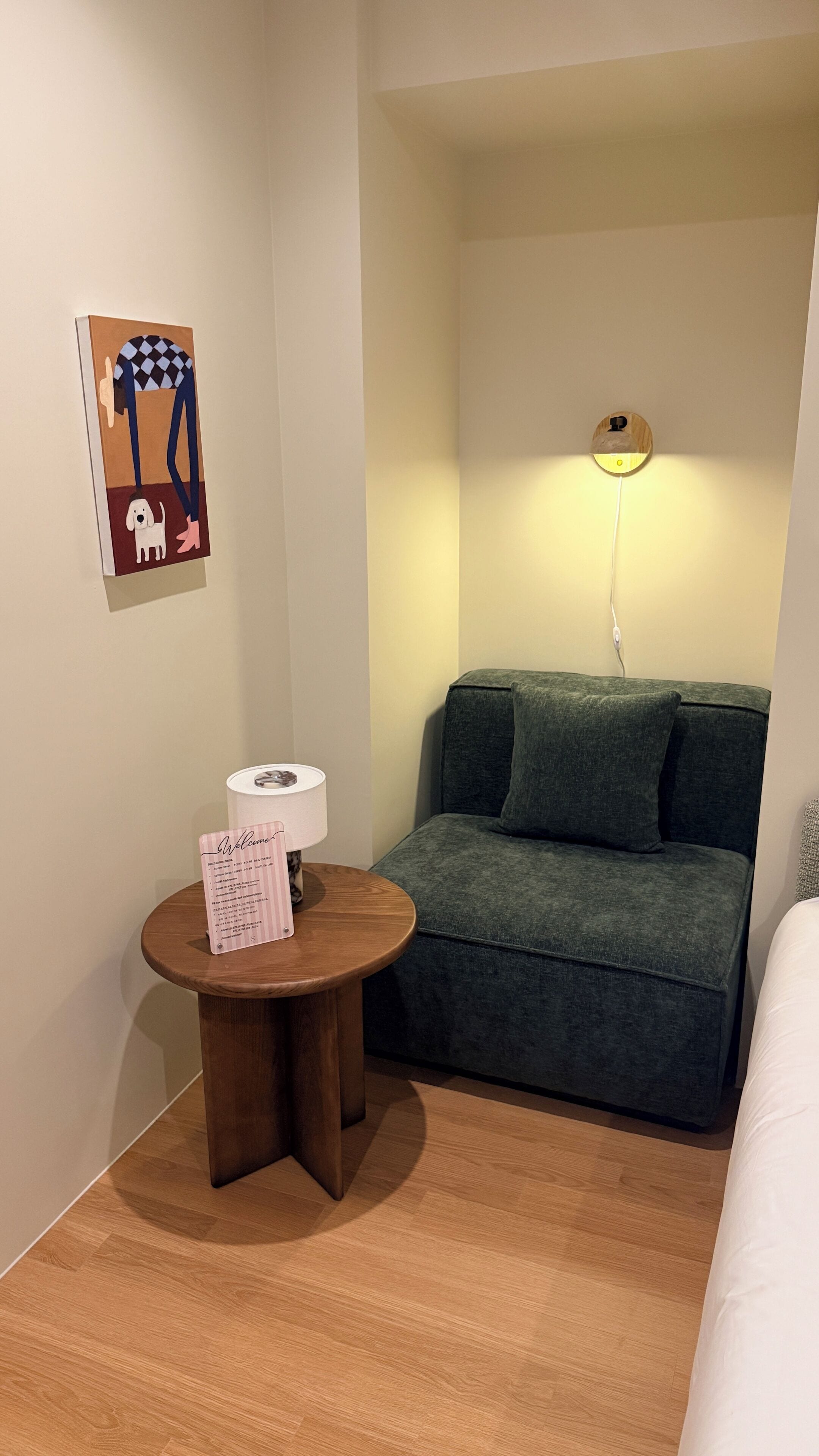 Una habitación con un sofá verde, una pequeña mesa de madera con un cartel de bienvenida, una lámpara y una lámpara de pared.