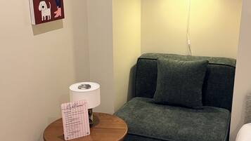 Una habitación con un sofá verde, una pequeña mesa de madera con un cartel de bienvenida, una lámpara y una lámpara de pared.