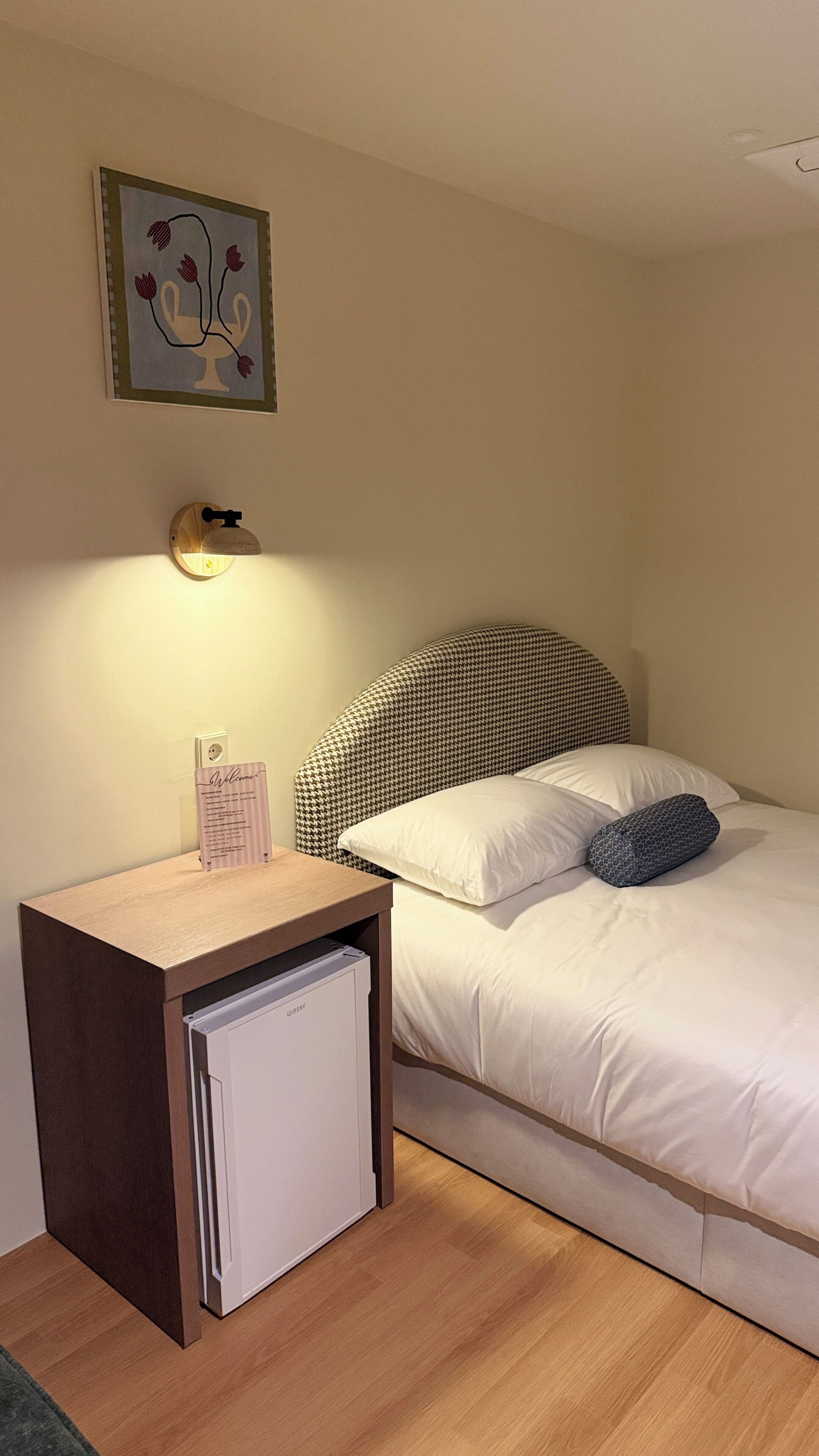 Una cama individual con cabecera a cuadros de gran perro, una mesita de noche con lámpara y un refrigerador pequeño.
