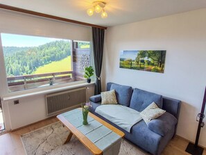 Een woonkamer met een bank, een salontafel, een raam met uitzicht op een bos, een radiator en een foto aan de muur.