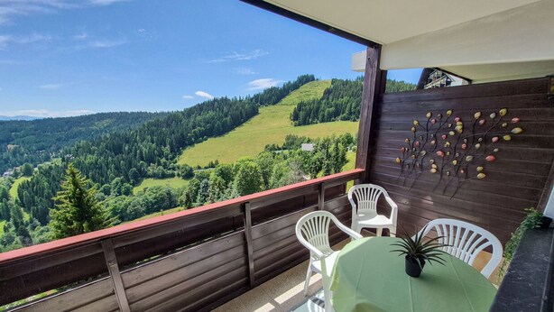 Un balcon donnant sur une vallée boisée, une clôture en bois et une table avec des chaises.