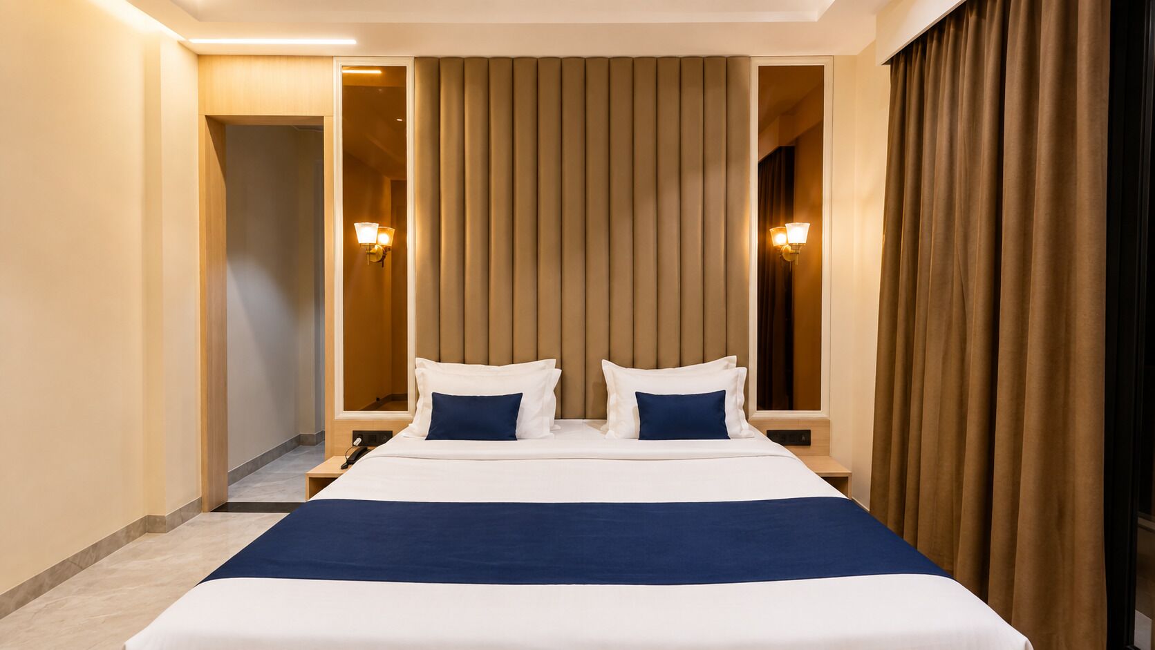 A hotel room with a large bed, a nightstand, a lamp, and a view of a corridor.