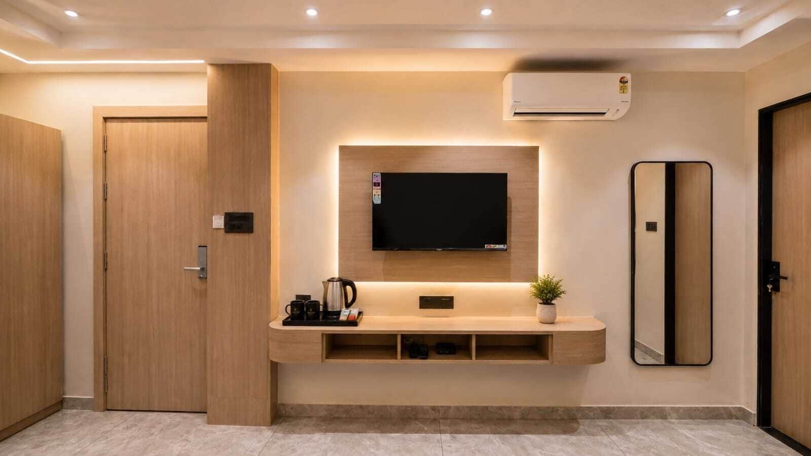A modern hotel room with a flat-screen TV mounted on a wooden wall, a coffee station, and a large mirror.