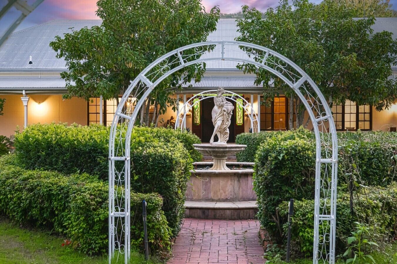 A garden with a statue under an arched structure, surrounded by well-maintained hedges and a pathway leading to a building with a metal roof.