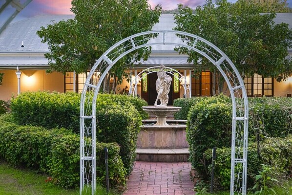 A garden with a statue under an arched structure, surrounded by well-maintained hedges and a pathway leading to a building with a metal roof.
