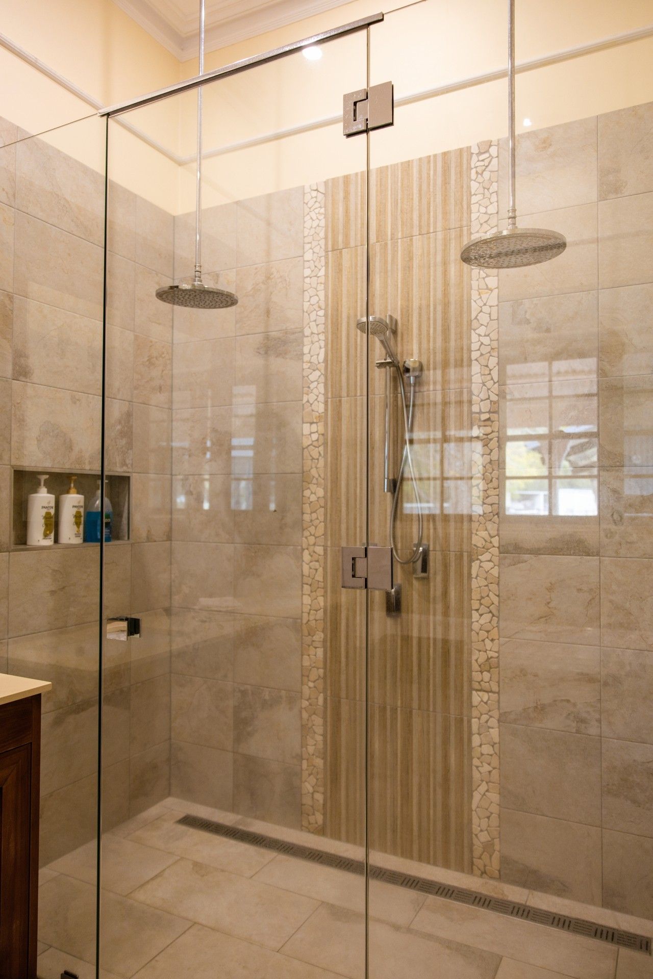 A glass-enclosed shower with a rain showerhead and handheld showerhead, a stone mosaic wall, and a small shelf with toiletries.