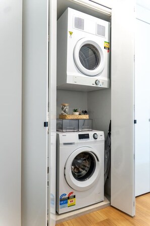 Ein kompakter Waschraum mit einer Waschmaschine und einem Trockner mit Frontlader, einer kleinen Ablage mit Dekoration und einer Tür mit einem Sicherheitshinweis.