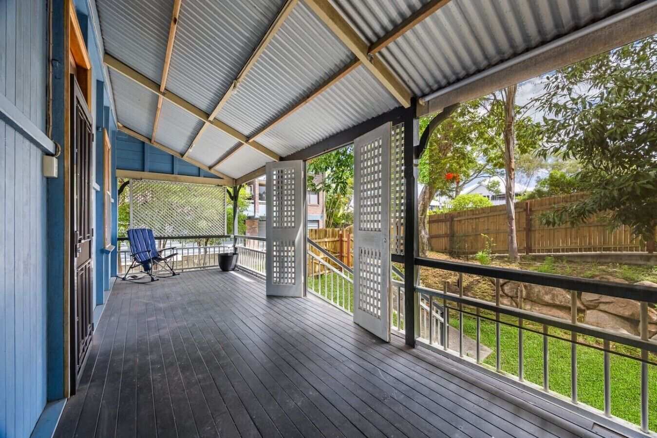 A covered outdoor area with a wooden deck, a blue chair, and a view of a fenced yard.