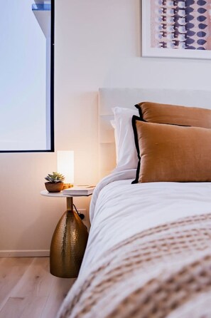 Ein ordentlich bezogenes Bett mit weißer und brauner Bettwäsche, ein kleiner Nachttisch mit Pflanze und ein an der Wand hängendes Kunstwerk.