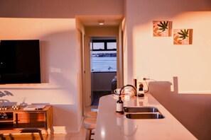 A modern kitchen with a large island, a dining area with a wooden table, and a view of a body of water through an open door. - Broadwater 2 bed 2 Bath 1 CarPK&Beach&Parks (Southport)