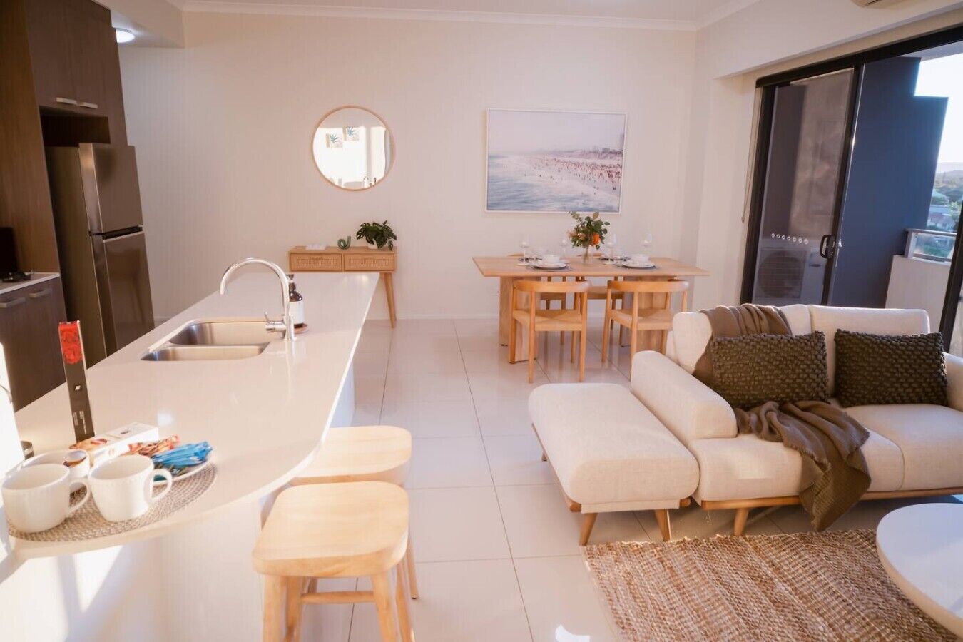 A modern kitchen with a central island, dining area with wooden chairs, and a sofa with a view of the outdoors.