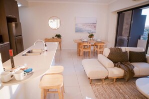 A modern kitchen with a central island, dining area with wooden chairs, and a sofa with a view of the outdoors. - Broadwater 2 bed 2 Bath 1 CarPK&Beach&Parks (Southport)