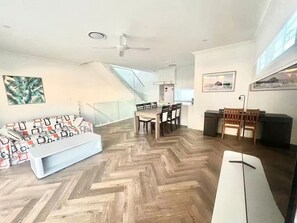 Amplia habitación con piso de espiga, zona de comedor con sillas y una mesa, sofá con cojines estampados, y una escalera que sube a un nivel superior.