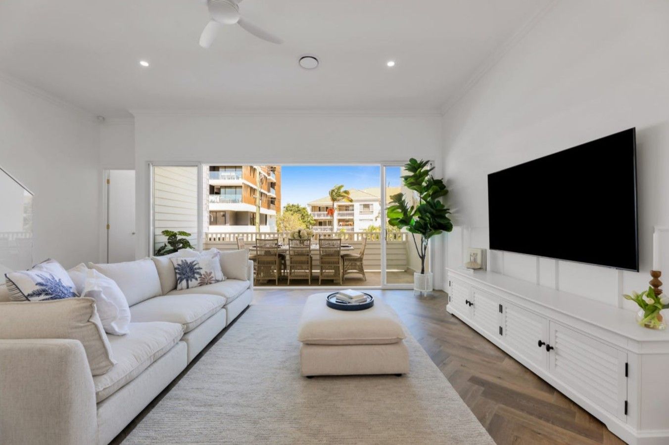 A modern living room with a sofa, ottoman, flat-screen TV, and a dining area with a view of buildings.