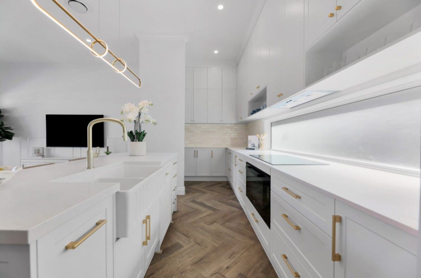 A modern kitchen with white cabinetry, gold handles, and a herringbone wood floor.
