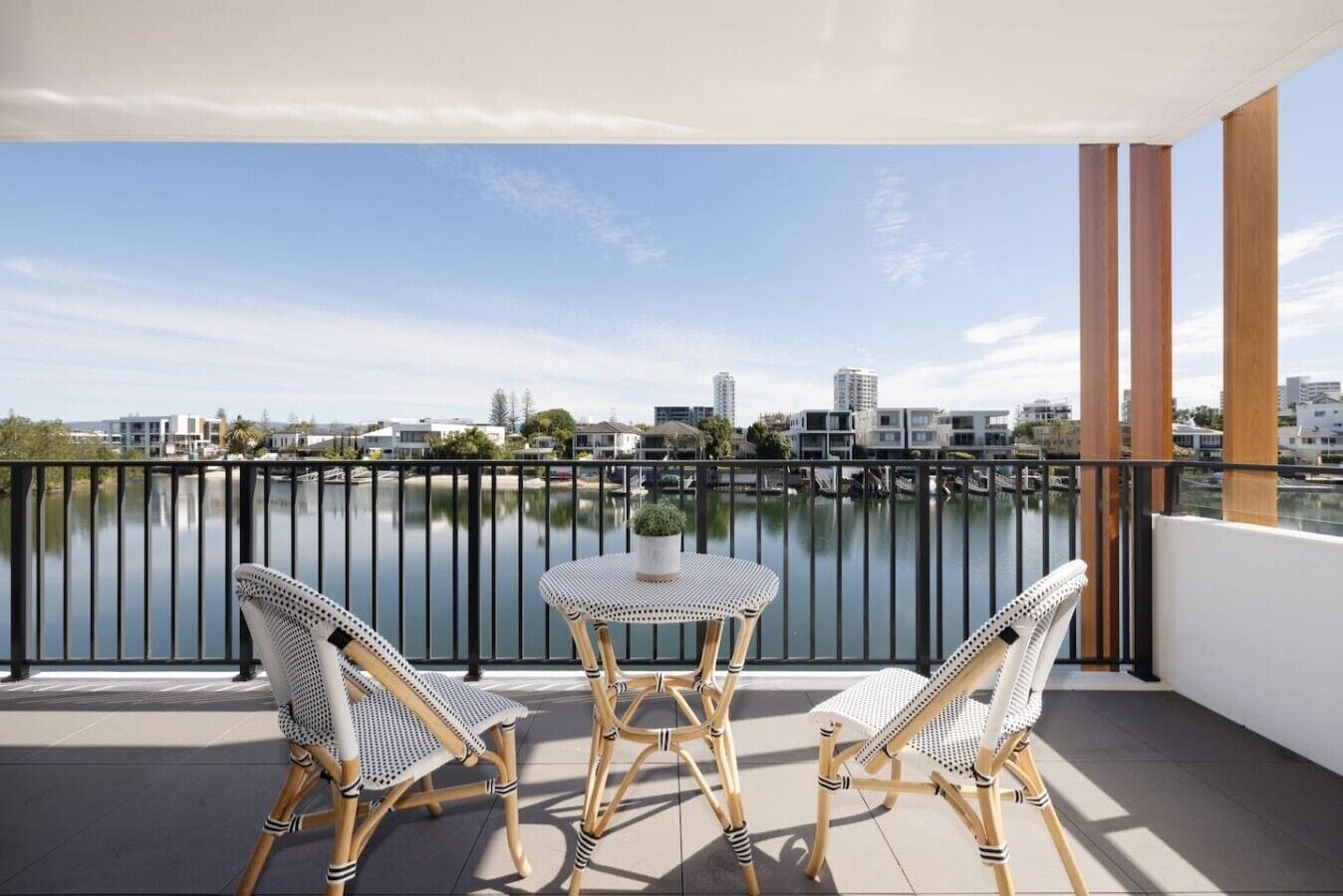 Een balkon met uitzicht op een rustig meer en gebouwen, voorzien van rieten stoelen en een klein tafeltje.