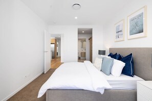 A modern bedroom with a large bed, a nightstand, and a headboard. There are framed pictures on the wall and a door leading to a bathroom. - Waterfront 2B 2Bath Pk,GYM,Pool&BBQ (Surfers Paradise)