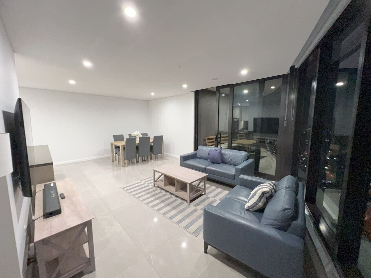 A modern living room with a sofa, coffee table, and TV stand. There is a dining area with chairs and a table.